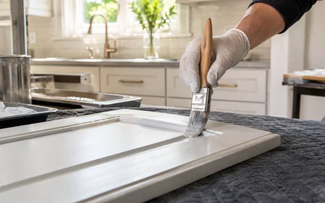 A gloved hand brushing white paint along the edge of a door on a padded table.